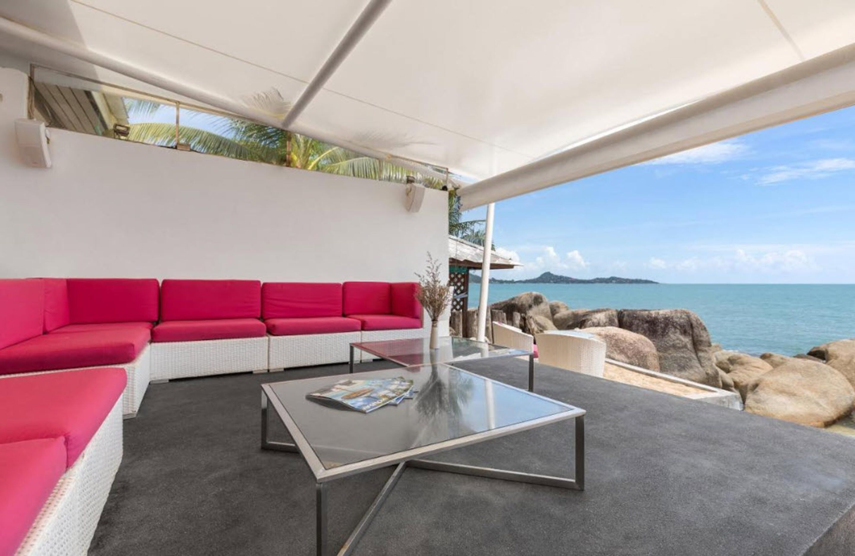 Un espacio exterior moderno con una zona de estar que incluye un sofá rojo y una mesa de centro de cristal, con vistas a un litoral rocoso y al mar.