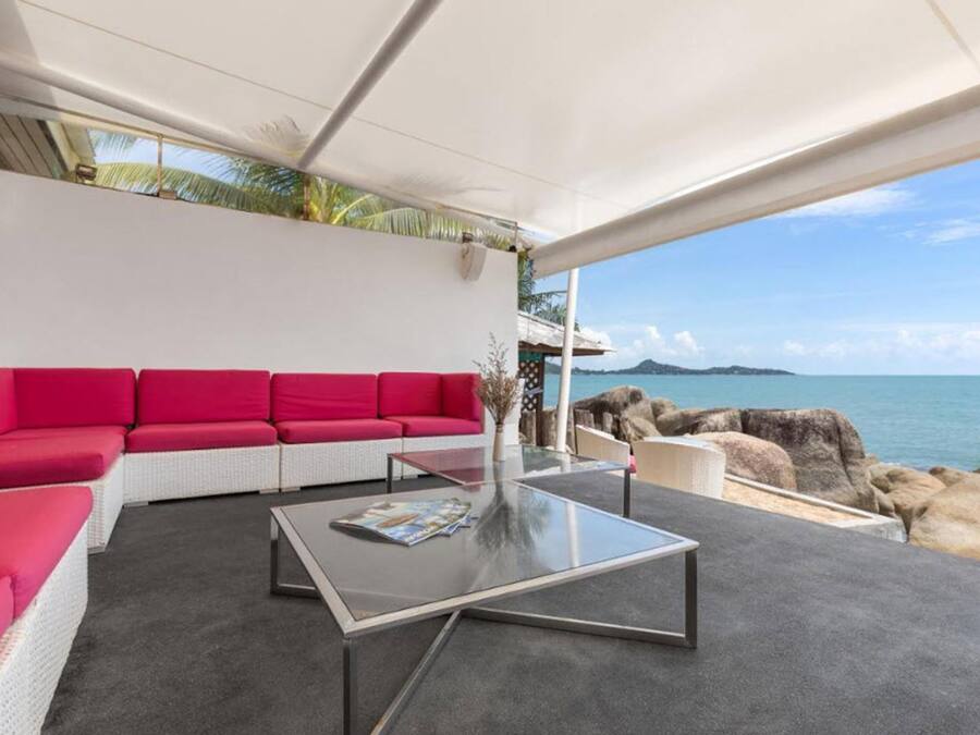 Un espacio exterior moderno con una zona de estar que incluye un sofá rojo y una mesa de centro de cristal, con vistas a un litoral rocoso y al mar.