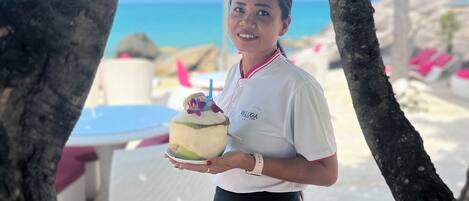 Una persona sosteniendo una bebida de coco con pajita y una flor encima, de pie bajo un árbol con una playa y sombrillas al fondo.