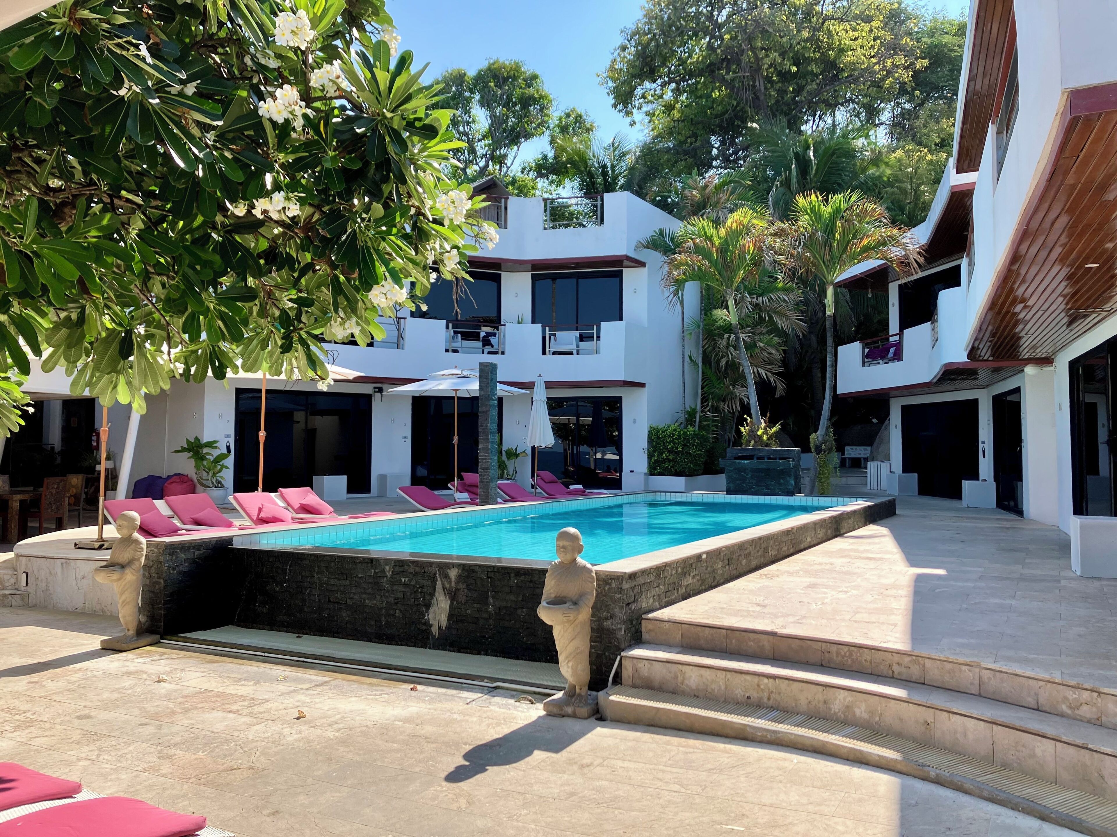 A modern pool area with a clear blue pool, lounge chairs with pink cushions, and a two-story building with balconies.