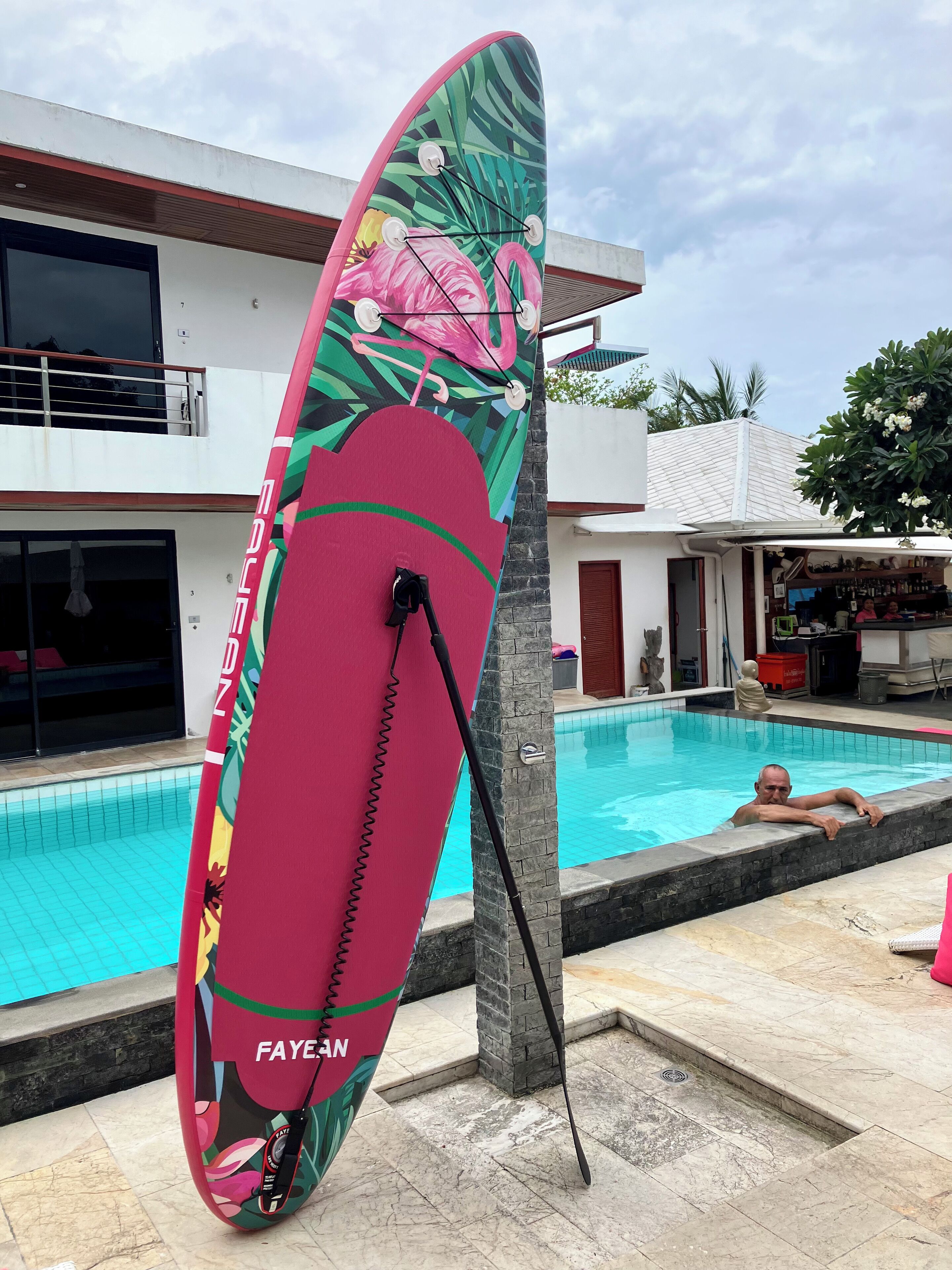 Zona de piscina con una tabla de surf colorida apoyada en un pilar, una piscina azul cristalina y un edificio con balcón al fondo.