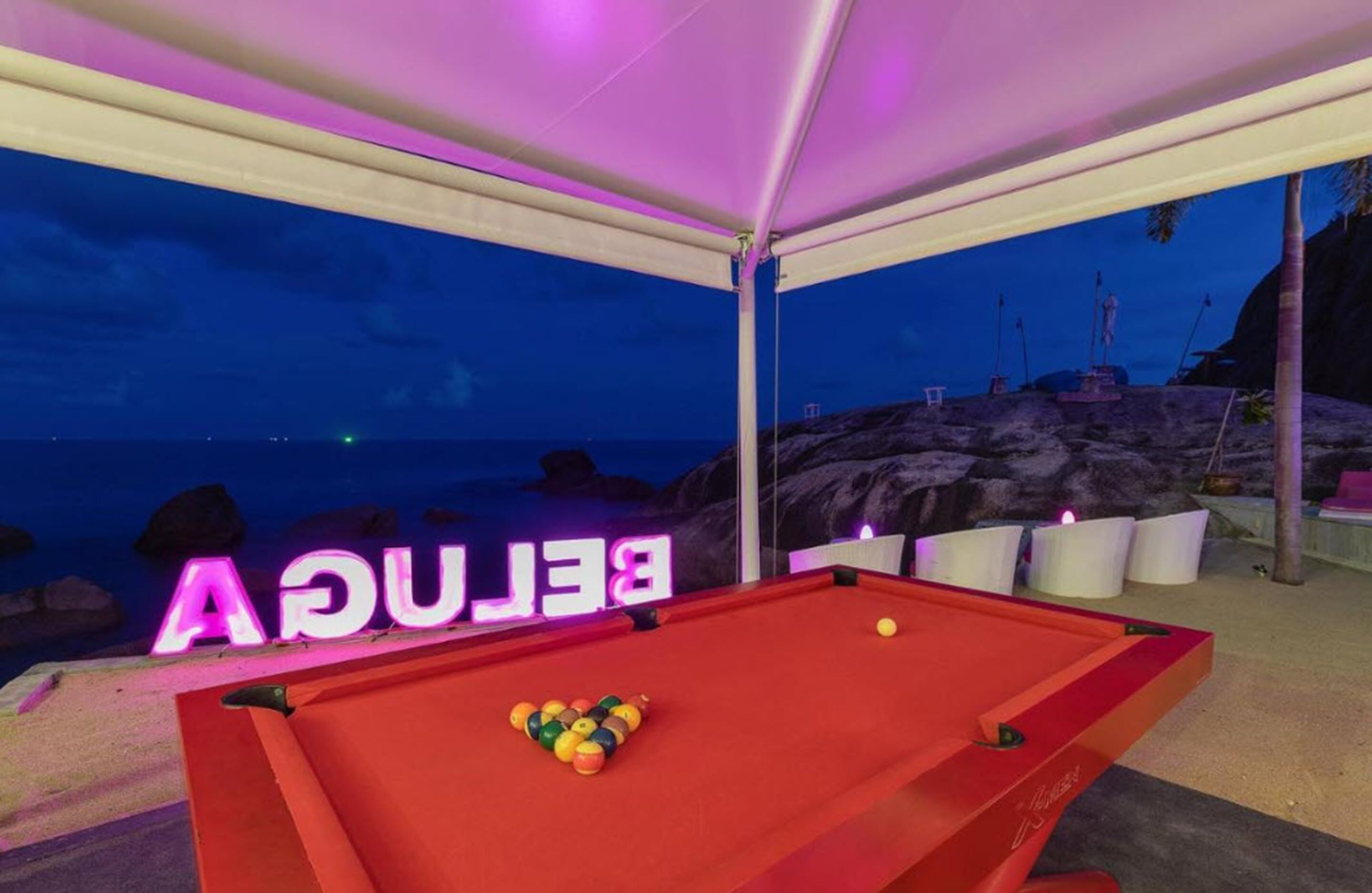 A pool table under a canopy with a neon sign reading "BELNAC" and a view of the beach at dusk.