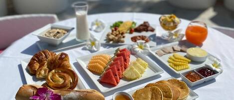 Una mesa blanca con un desayuno que incluye pasteles, frutas y condimentos, con vista a una playa con rocas y el mar.