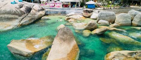 A clear pool with large rocks, surrounded by houses and palm trees.