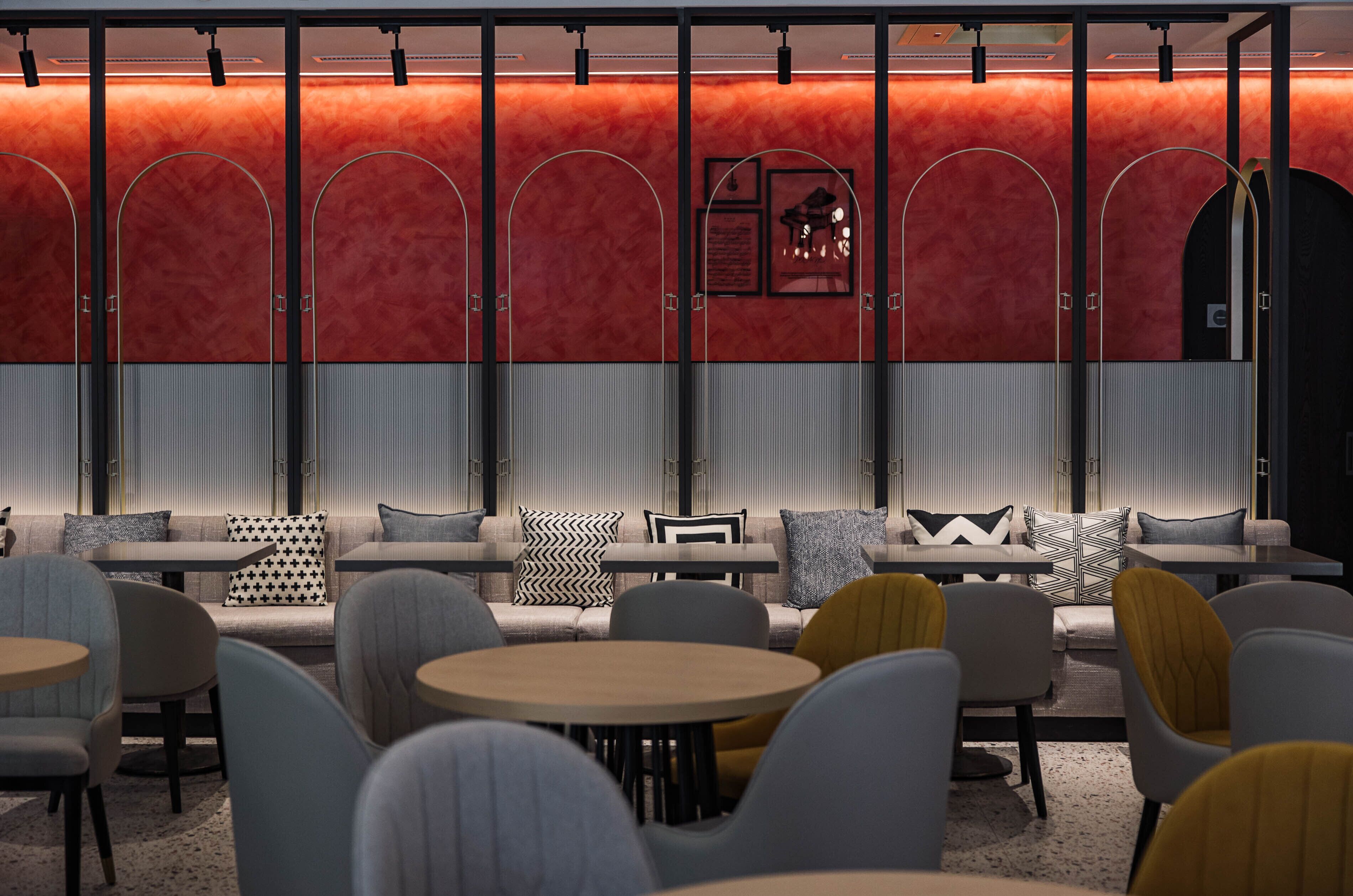 A modern lounge area with seating and tables, featuring a red wall with a framed artwork and a black door.