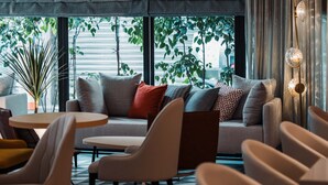 A modern lounge area with a sofa, chairs, and a small table. Large windows provide a view of greenery outside. - OLAH Poshtel - Taichung Station (Taichung)