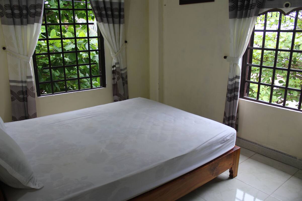 A room with a bed, two windows with curtains, and a view of greenery outside.