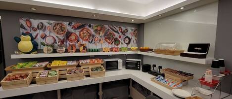 Una cocina moderna con microondas, tostadora y cafetera sobre la encimera. Hay cajas de madera con aperitivos y una cesta de fruta. Un estante muestra una variedad de alimentos y una caja de Toronton.