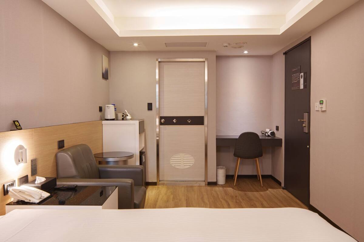 A hotel room with a bed, a desk, a chair, a small table, a coffee maker, and a door.
