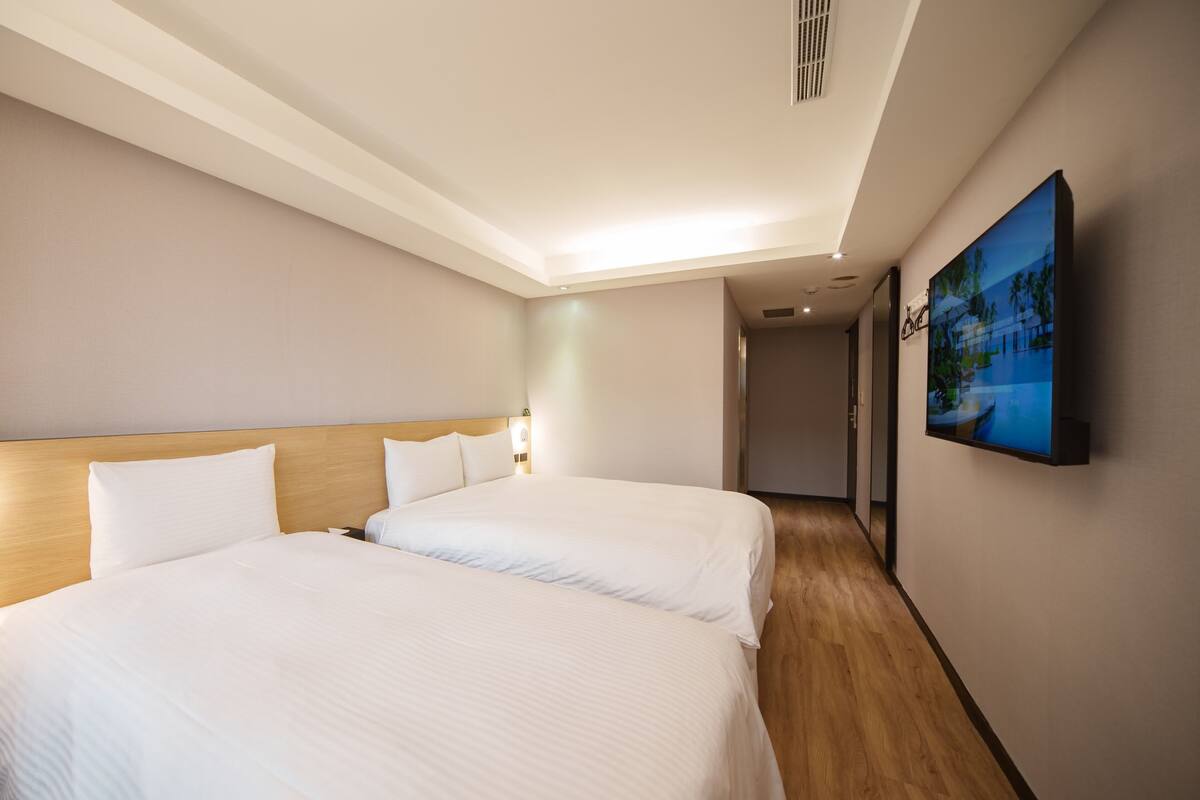 A hotel room with two beds, a wooden headboard, a flat-screen TV on the wall, and a hallway leading to another room.