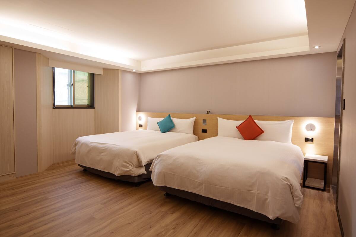 A hotel room with two beds, wooden flooring, a window, and bedside lamps.