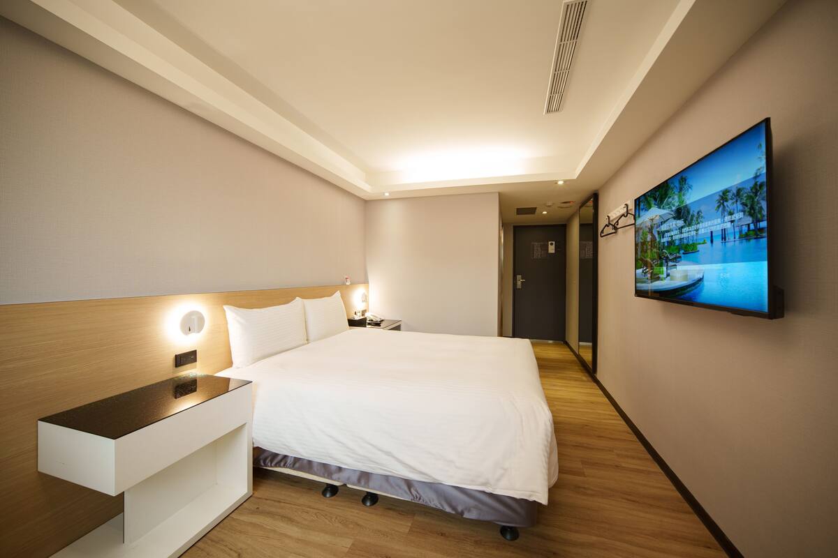A modern hotel room with a large bed, bedside table, and a flat-screen TV mounted on the wall.