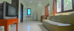 A living room with a television on a wooden stand, a sofa, a window with curtains, and a refrigerator in the background.