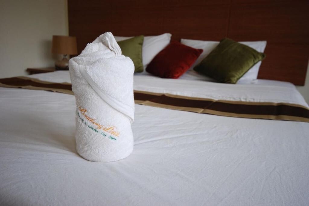 Una cama bien hecha con una toalla blanca enrollada en el centro, un cabecero marrón y varios cojines en rojo, verde y blanco.