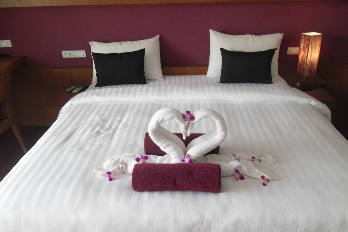 A hotel room with a neatly made bed featuring a swan-shaped towel arrangement and purple flowers.