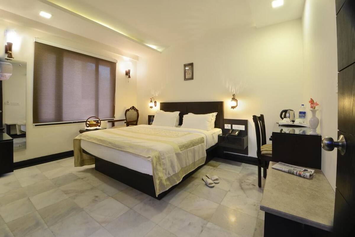 Habitación de hotel con una cama grande, mesitas de noche, un escritorio, una silla, una ventana con persianas y una puerta.