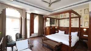 Una cama con dosel de madera, un banco, una mesa y una silla en una habitación con paredes estampadas y un ventilador de techo.