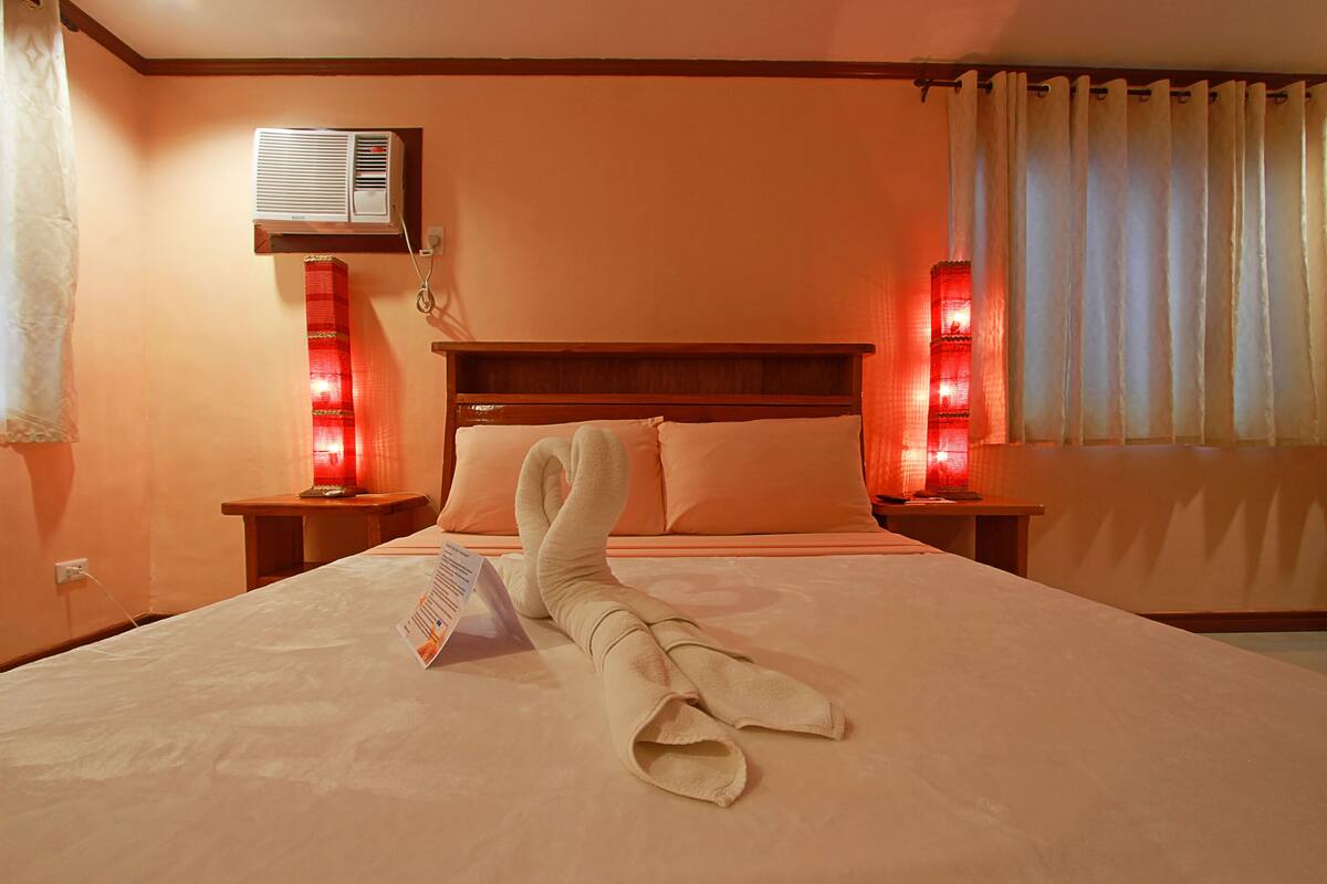 A hotel room with a neatly made bed, a bedside table, a nightstand, a wall-mounted air conditioner, and a window with curtains.