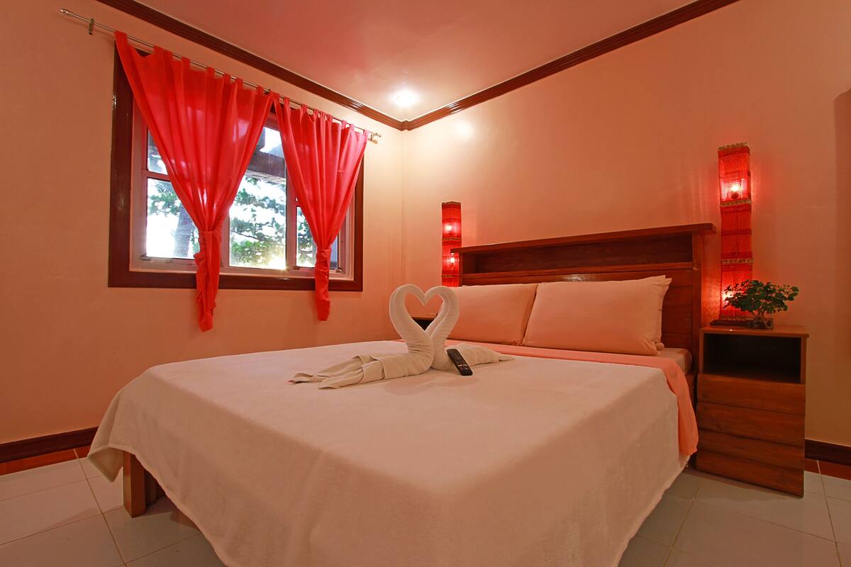 A neatly made bed with a heart-shaped towel arrangement, a bedside table with a plant, and a window with red curtains.