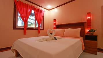 A neatly made bed with a heart-shaped towel arrangement, a bedside table with a plant, and a window with red curtains.