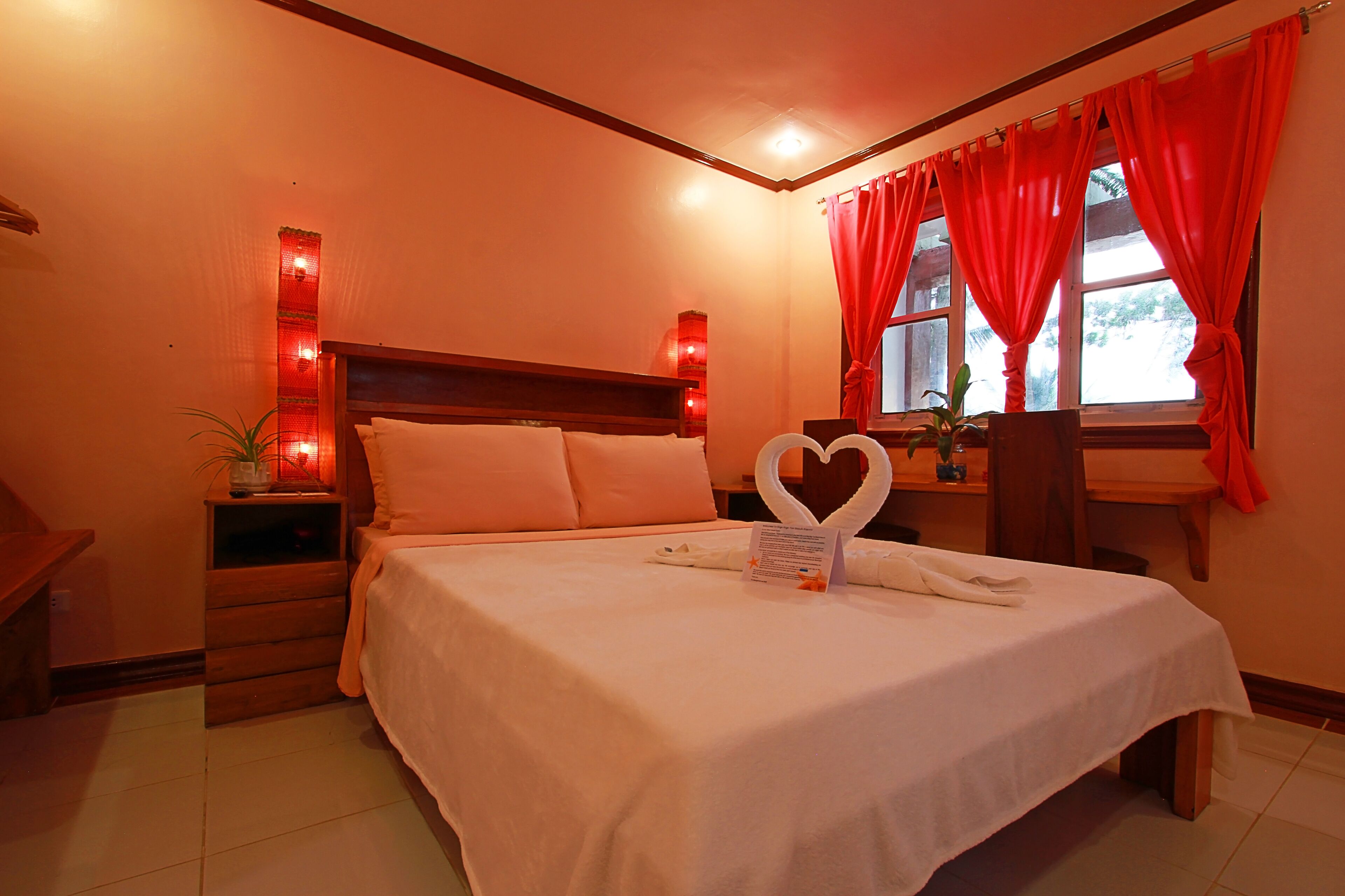 Ein Hotelzimmer mit Bett, Nachttisch, Fenster mit roten Vorhängen und einer Wandleuchte.