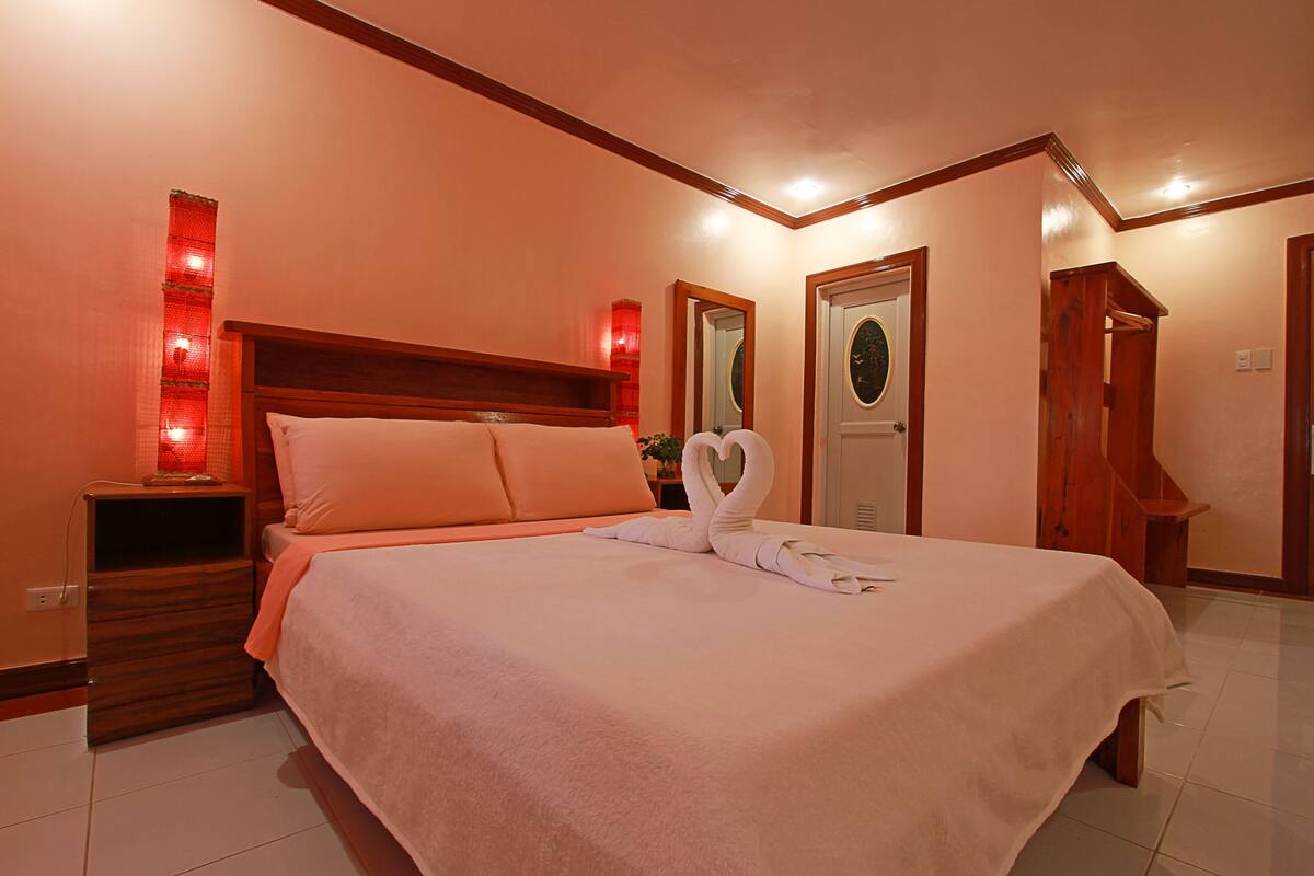 A neatly made bed with a heart-shaped towel arrangement, a wooden headboard, and a nightstand with a red lamp.