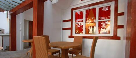 A room with a round table and four wicker chairs, a window with red curtains, and a wooden ceiling structure.