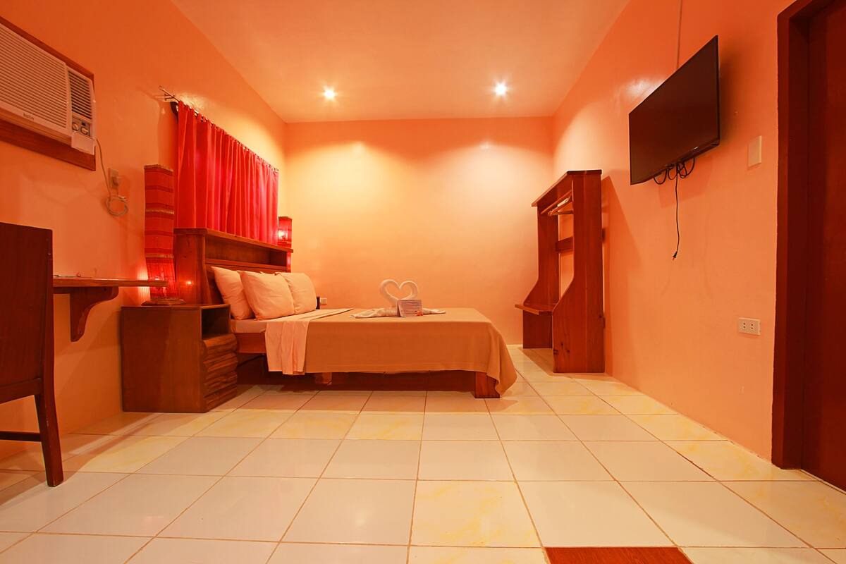 A bedroom with a bed, a bedside table, a TV mounted on the wall, and a wooden wardrobe.