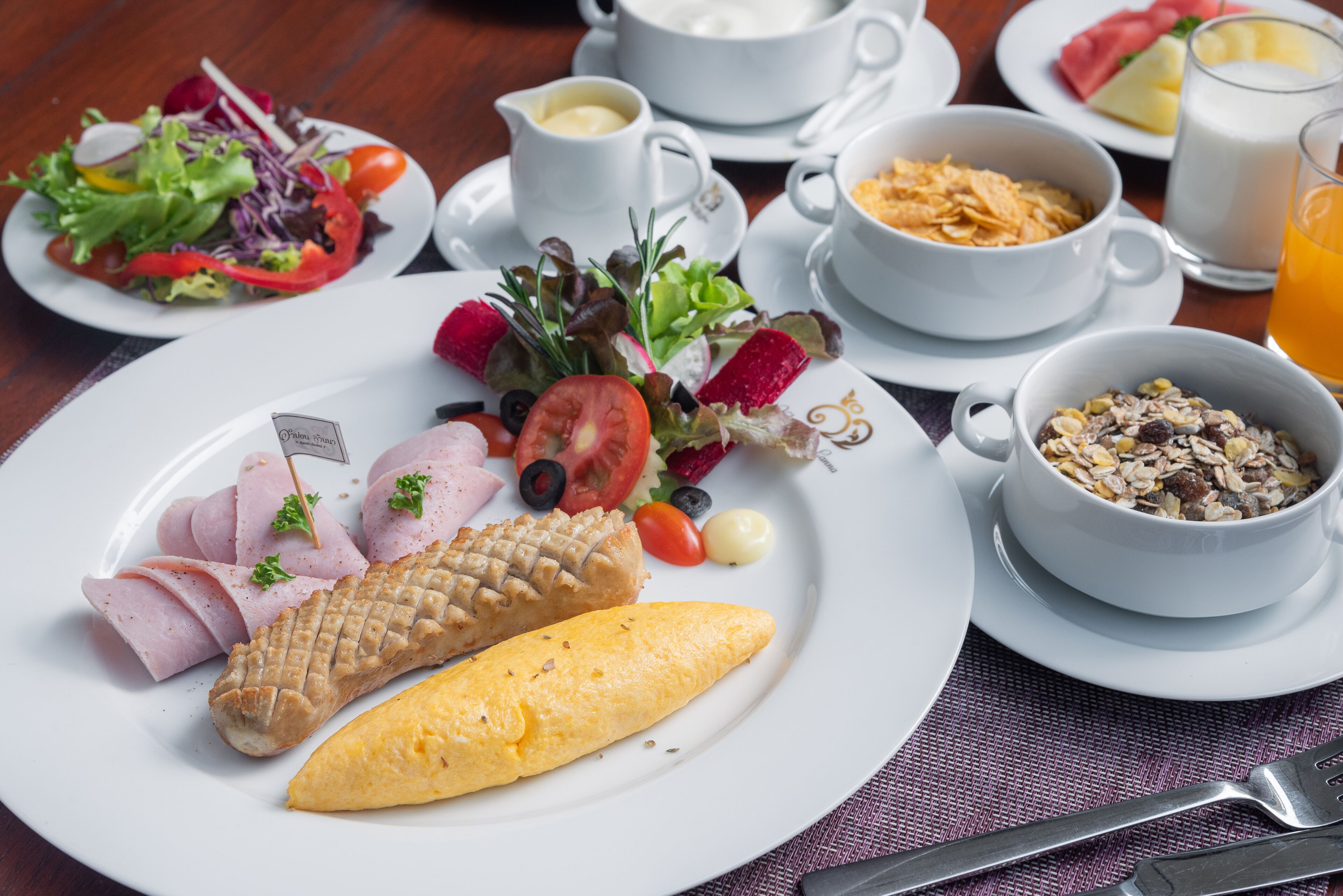A breakfast setting with plates of ham, eggs, and a pastry, accompanied by a salad and a bowl of cereal.