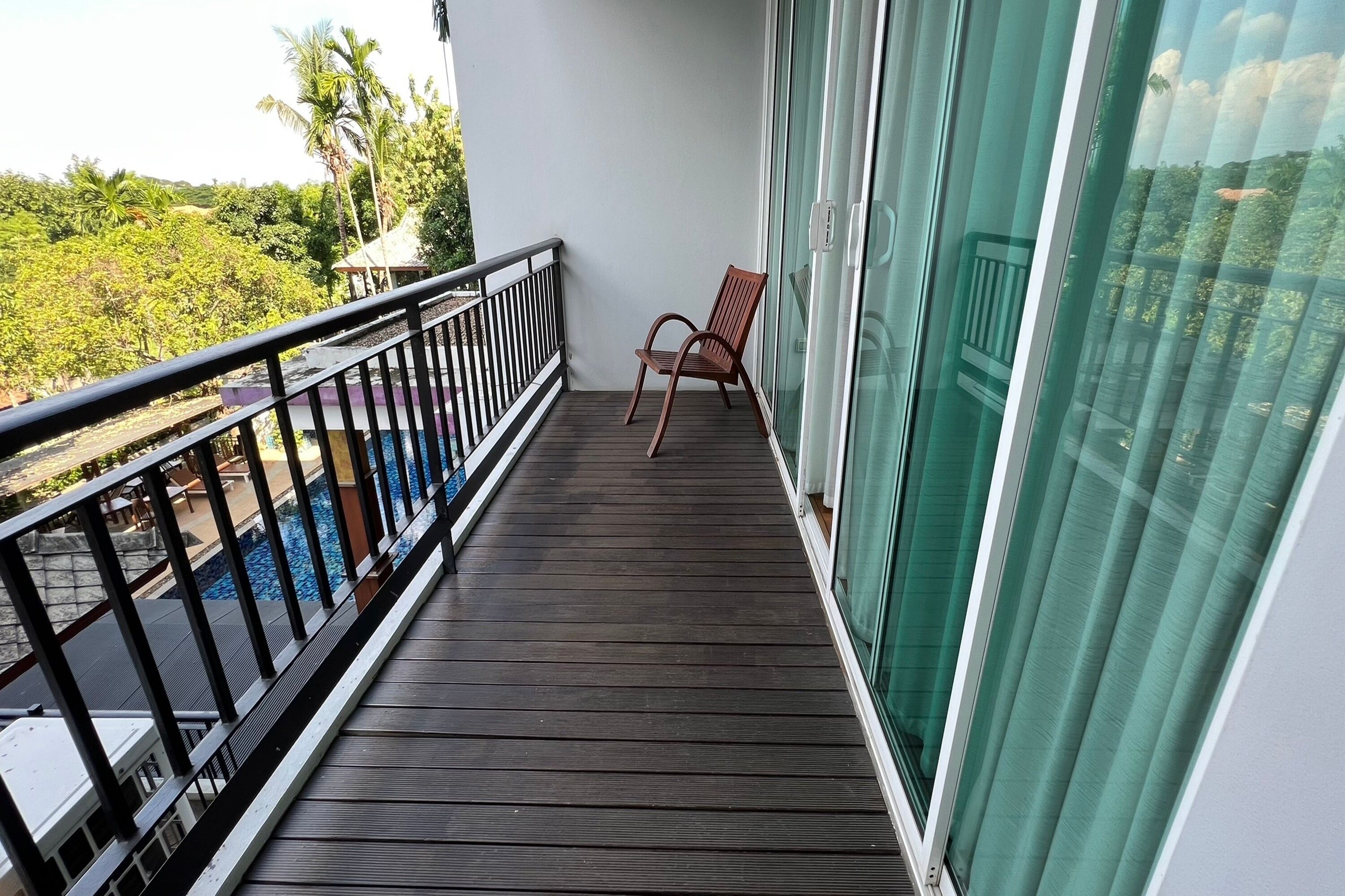 Un balcón con piso de madera, una silla y vista a una piscina y follaje.