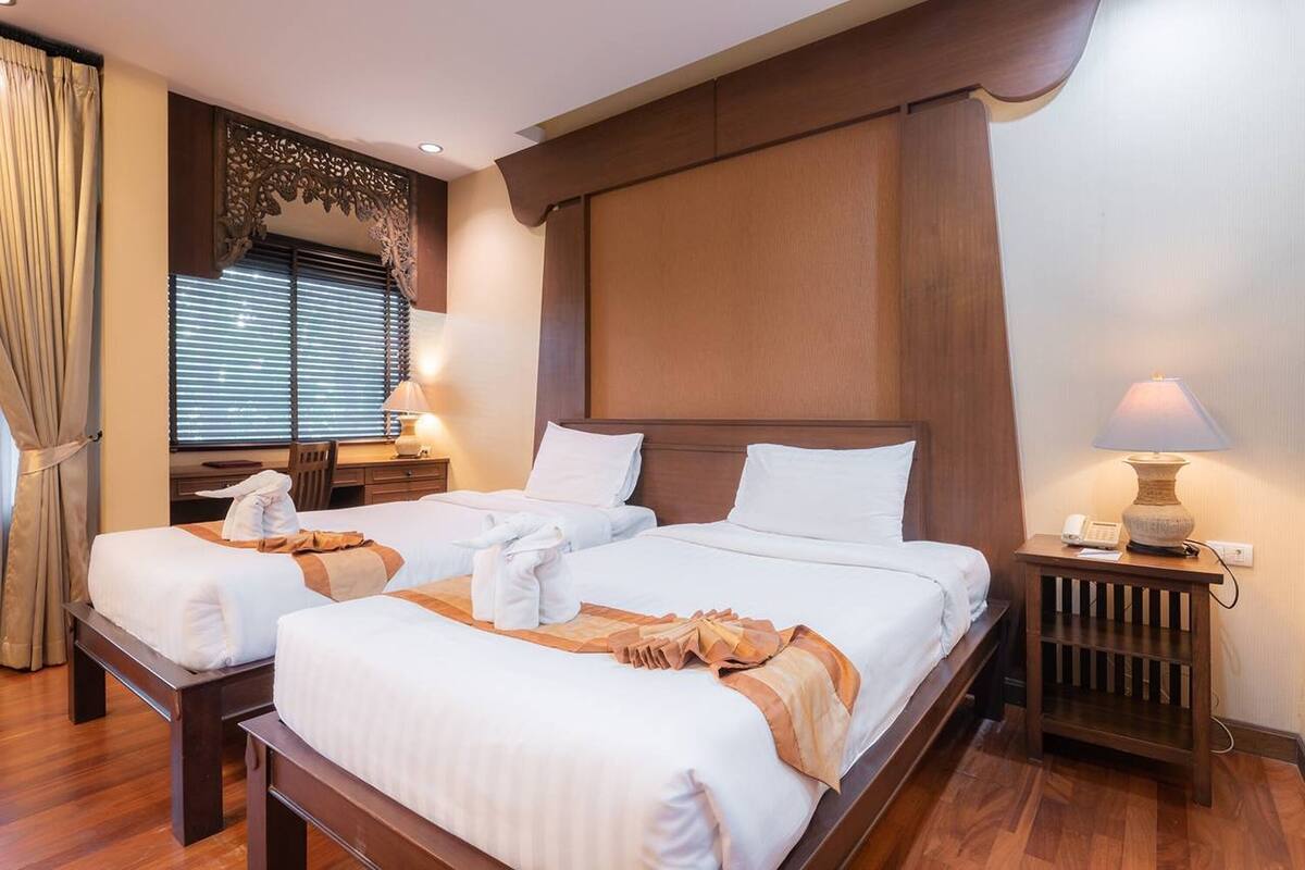 Hotelkamer met twee bedden, houten hoofdborden, een raam met zonwering, een bureau en een nachtkastje met een lamp.