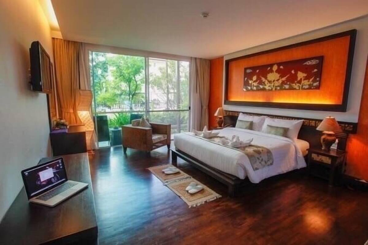 A hotel room with a bed, a desk with a laptop, a large window, and a painting on the wall.