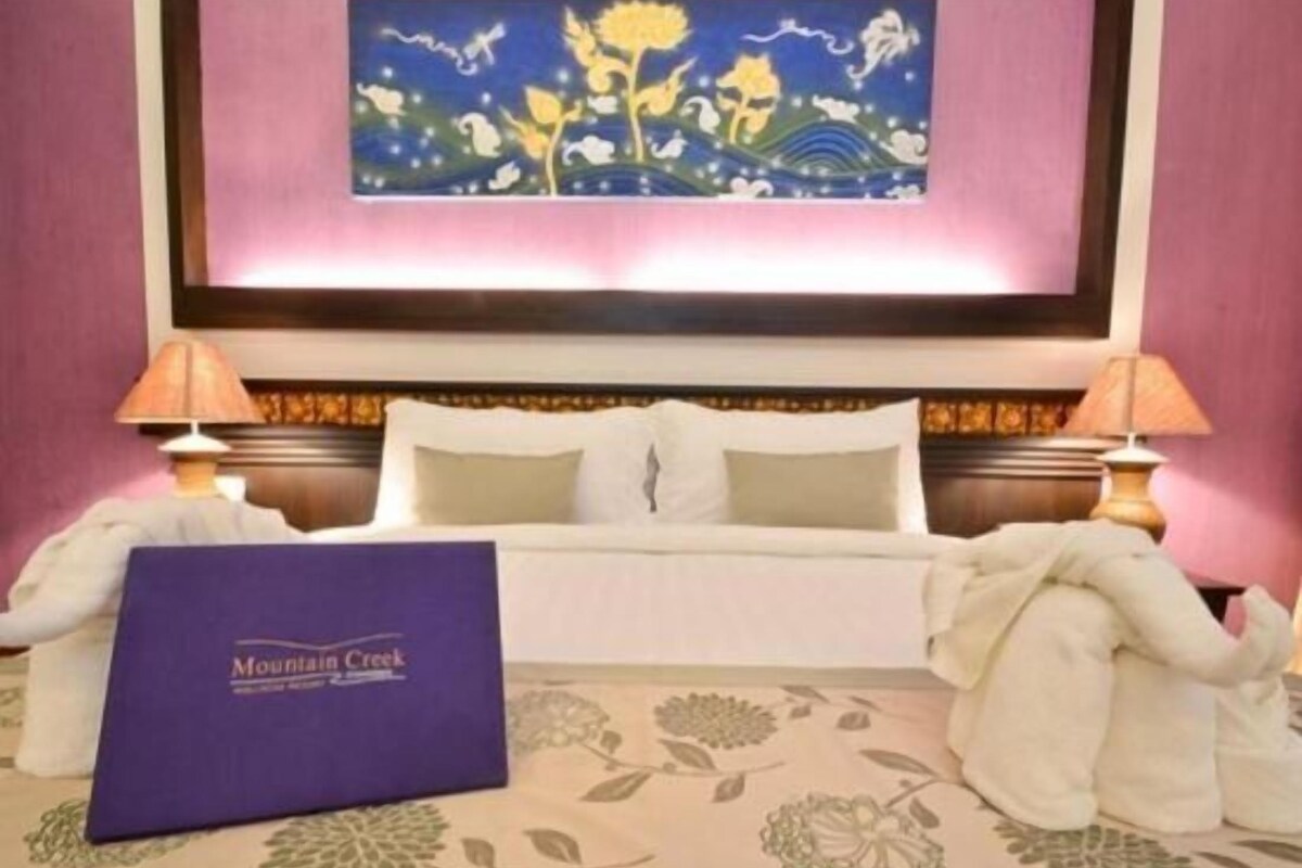 A hotel room with a bed, two bedside lamps, a painting on the wall, and a purple folder labeled "Mountain Creek" on the bed.