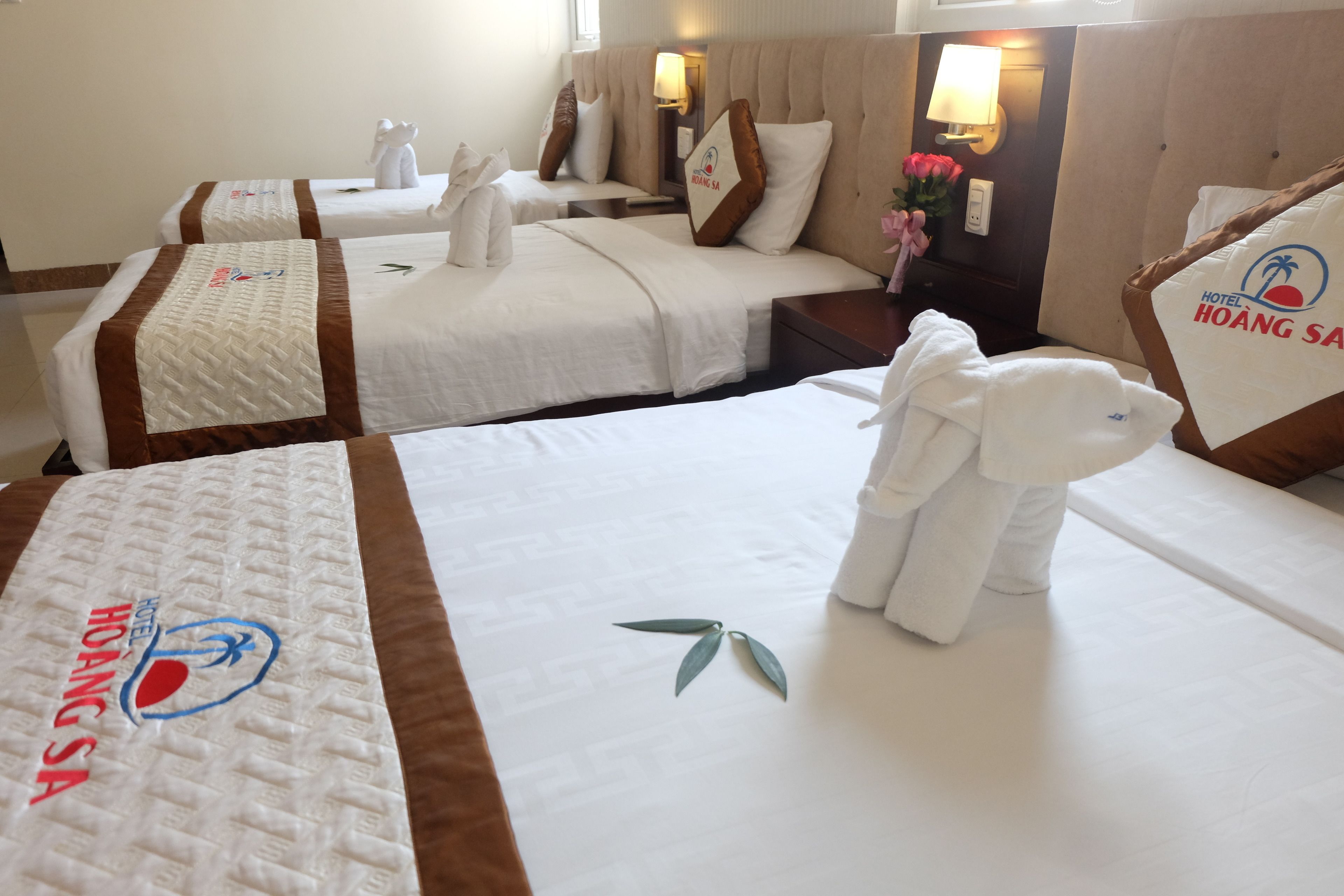 Une chambre d’hôtel avec deux lits, chacun équipé d’un oreiller et d’une serviette. On y trouve une table de chevet avec une lampe et un vase à fleurs. La chambre est décorée dans des tons neutres, avec des touches de beige et de marron.