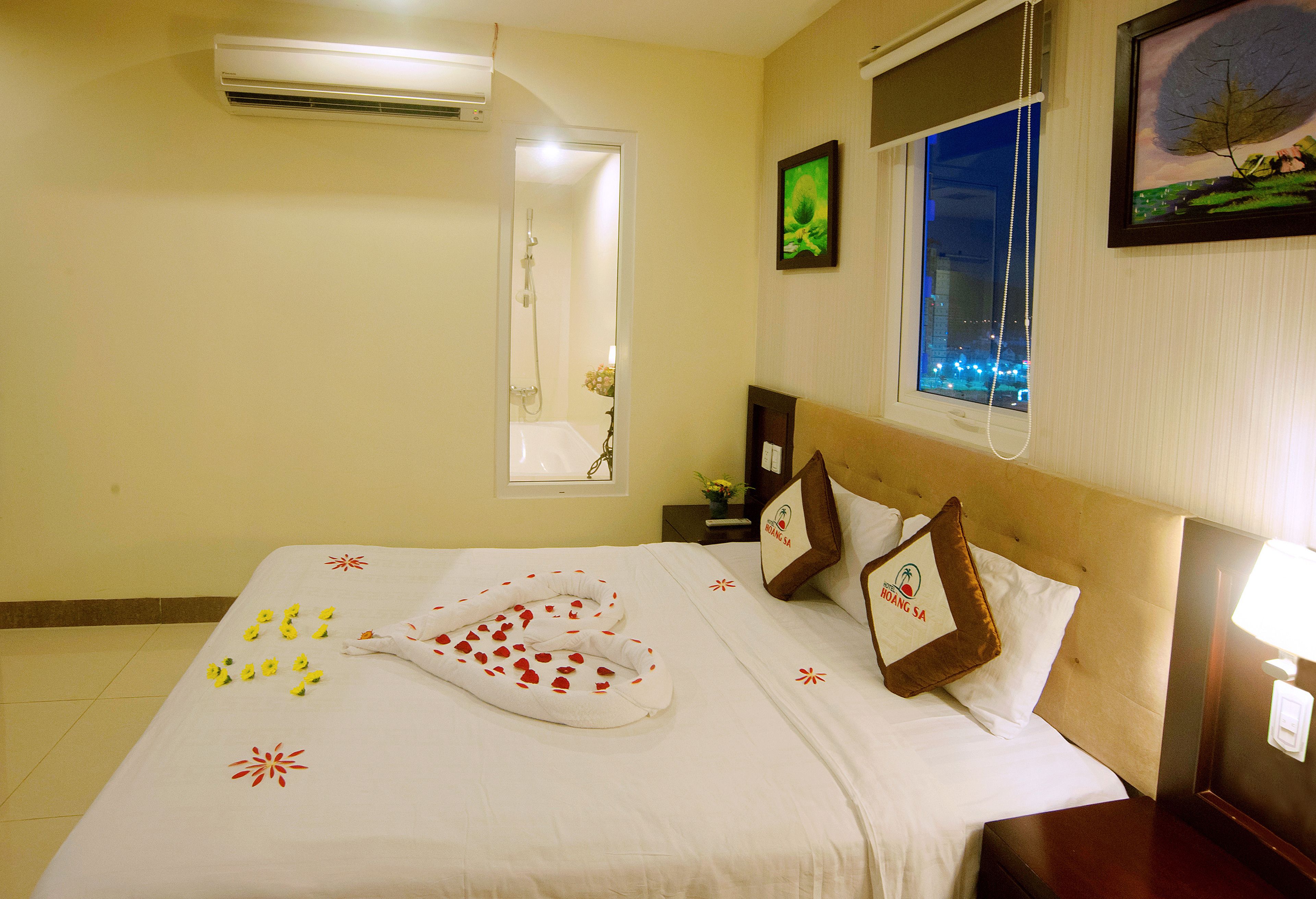 A hotel room with a bed, a window with a city view, a wall-mounted air conditioner, and a nightstand with a lamp.