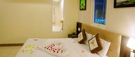 A hotel room with a bed, a window with a city view, a wall-mounted air conditioner, and a nightstand with a lamp.