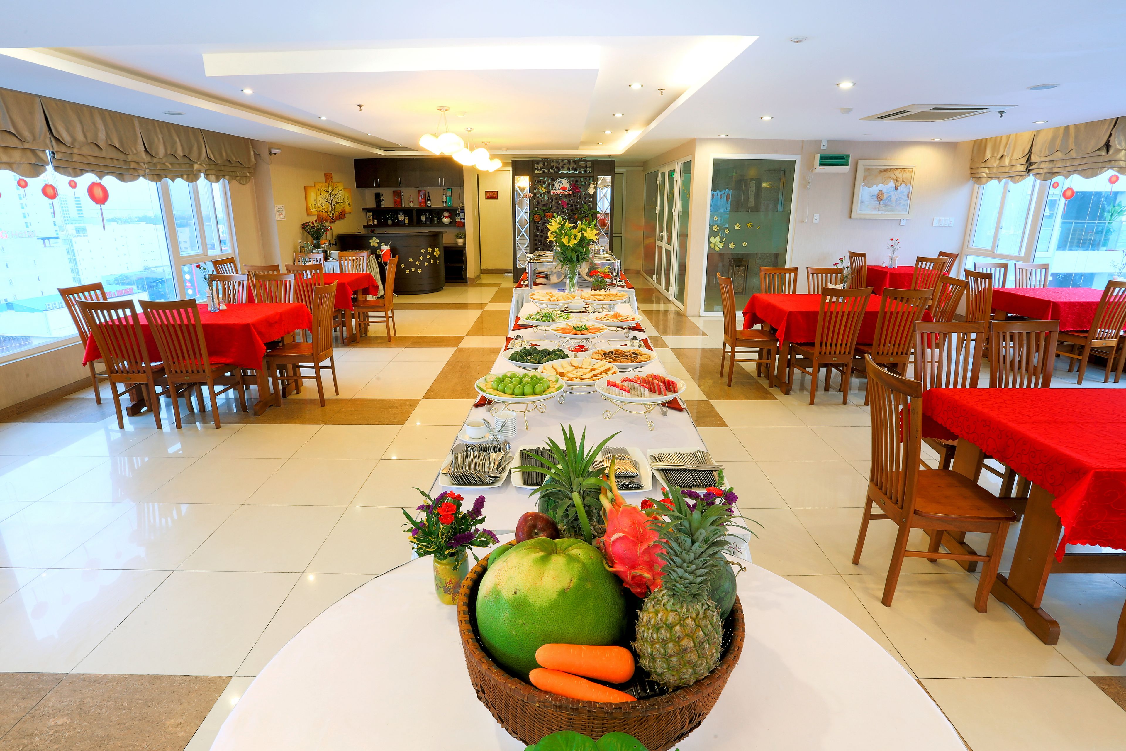 Un comedor con varias mesas cubiertas con manteles rojos, dispuesto en una sala amplia con techo alto y ventanas grandes.