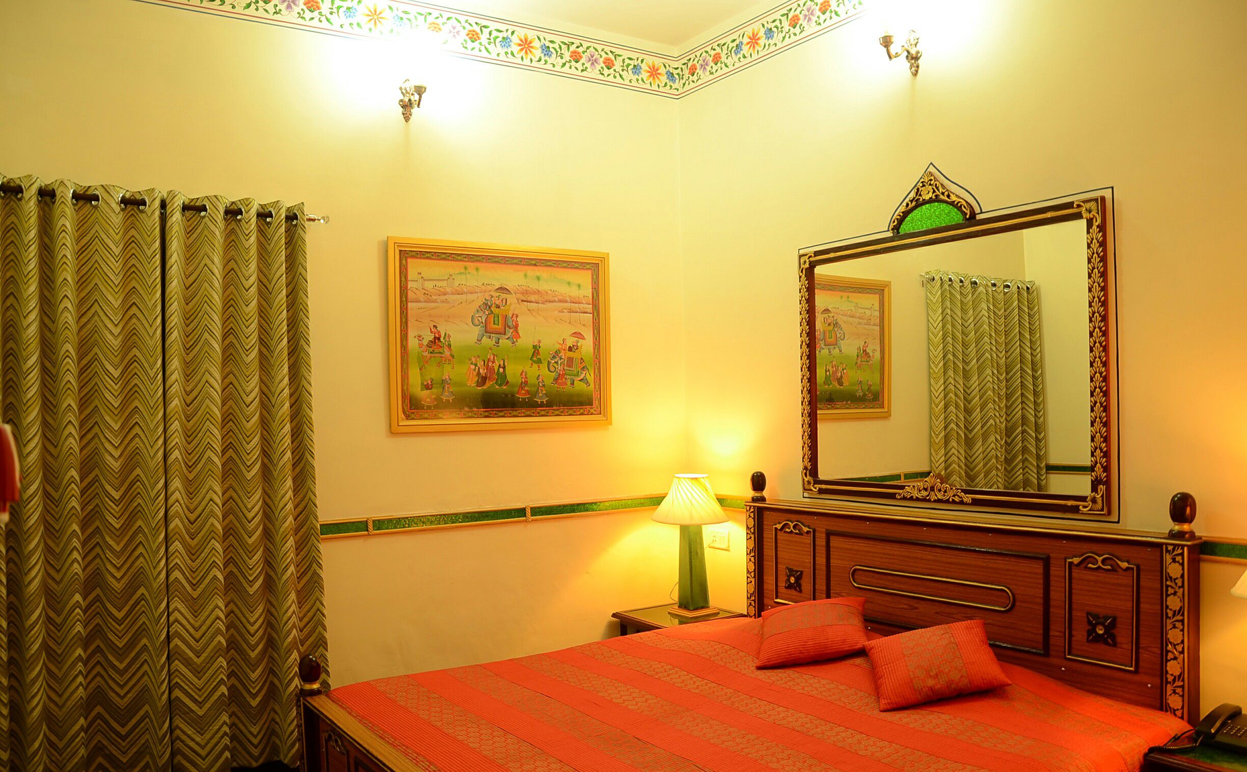 Habitación de hotel con cama, mesita de noche, lámpara, espejo, pared con un borde floral y cortina.