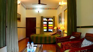 A hotel room with a bed, two chairs, a table with water bottles, a ceiling fan, and a window with colorful stained glass.