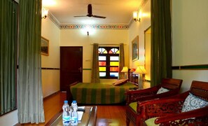 Quarto suíte com cama, duas cadeiras, mesa com garrafas de água, ventilador de teto e janela com cortinas.