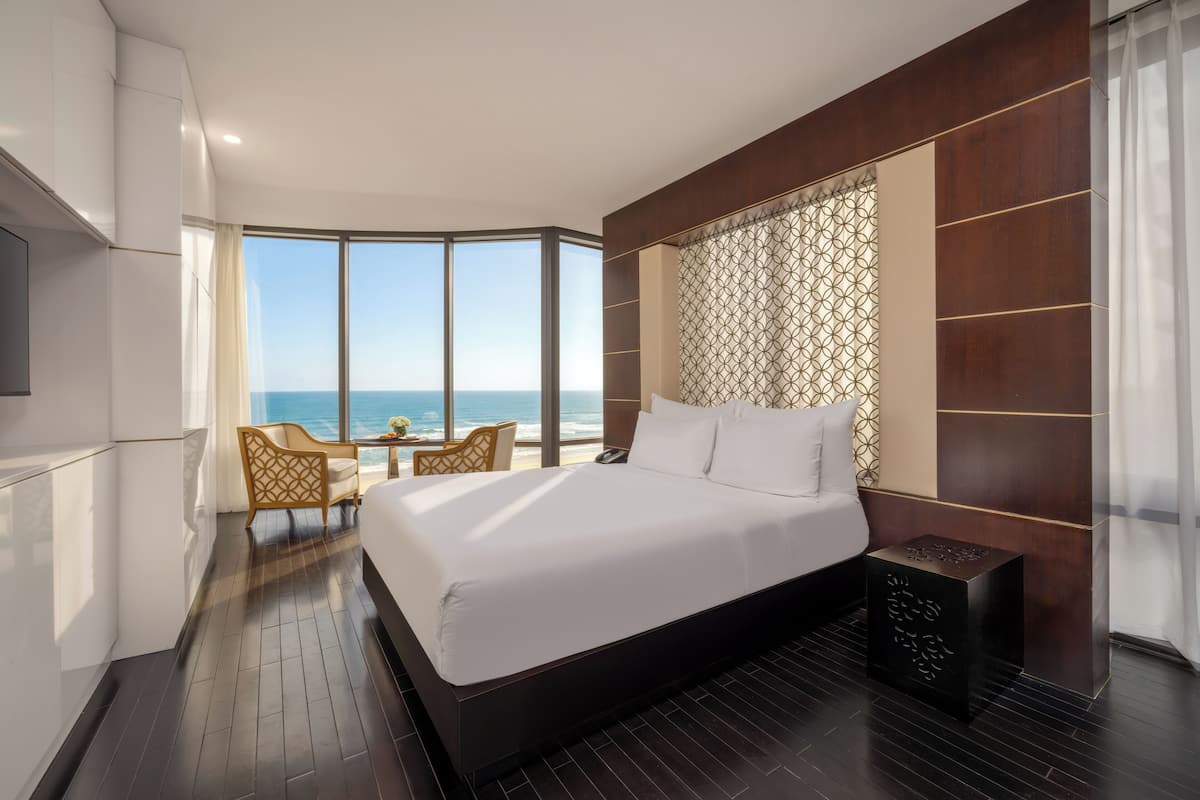 Une chambre d’hôtel moderne dotée d’un grand lit, d’un coin salon avec vue sur l’océan et d’une grande fenêtre.