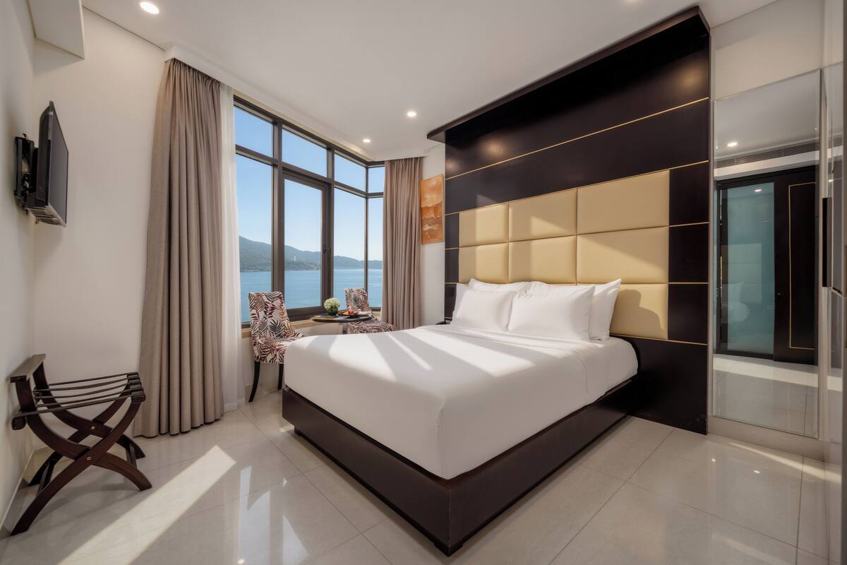 Une chambre d’hôtel moderne avec un grand lit, une télévision, une fenêtre donnant sur la mer et une petite table avec une chaise.