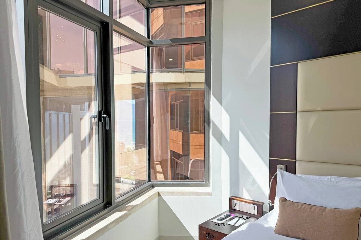 Une chambre d’hôtel moderne dotée d’une grande fenêtre, d’un lit avec une literie blanche, d’une table de chevet et offrant une vue sur le balcon extérieur.