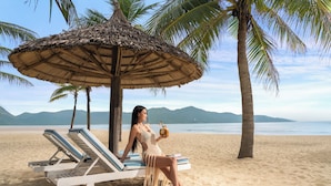 A beach scene with a woman sitting on a lounge chair under a thatched umbrella, palm trees, and a view of the sea and mountains.
