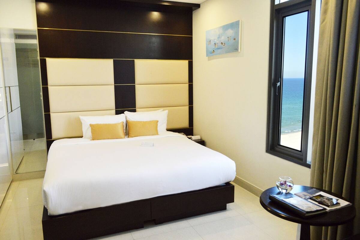 Une chambre d’hôtel avec un grand lit, une petite table de chevet, une fenêtre donnant sur la mer et une tête de lit moderne.