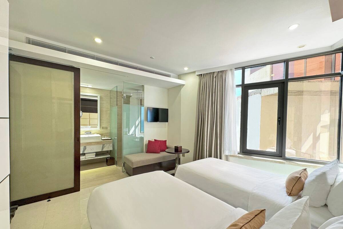 Une chambre d’hôtel avec deux lits, une télévision et une salle de bain dotée d’une paroi en verre, ainsi qu’une grande fenêtre.