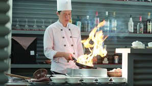 A chef cooking with flames in a kitchen with shelves of bottles and glasses in the background. - Diamond D'Angkor Boutique (Siem Reap)