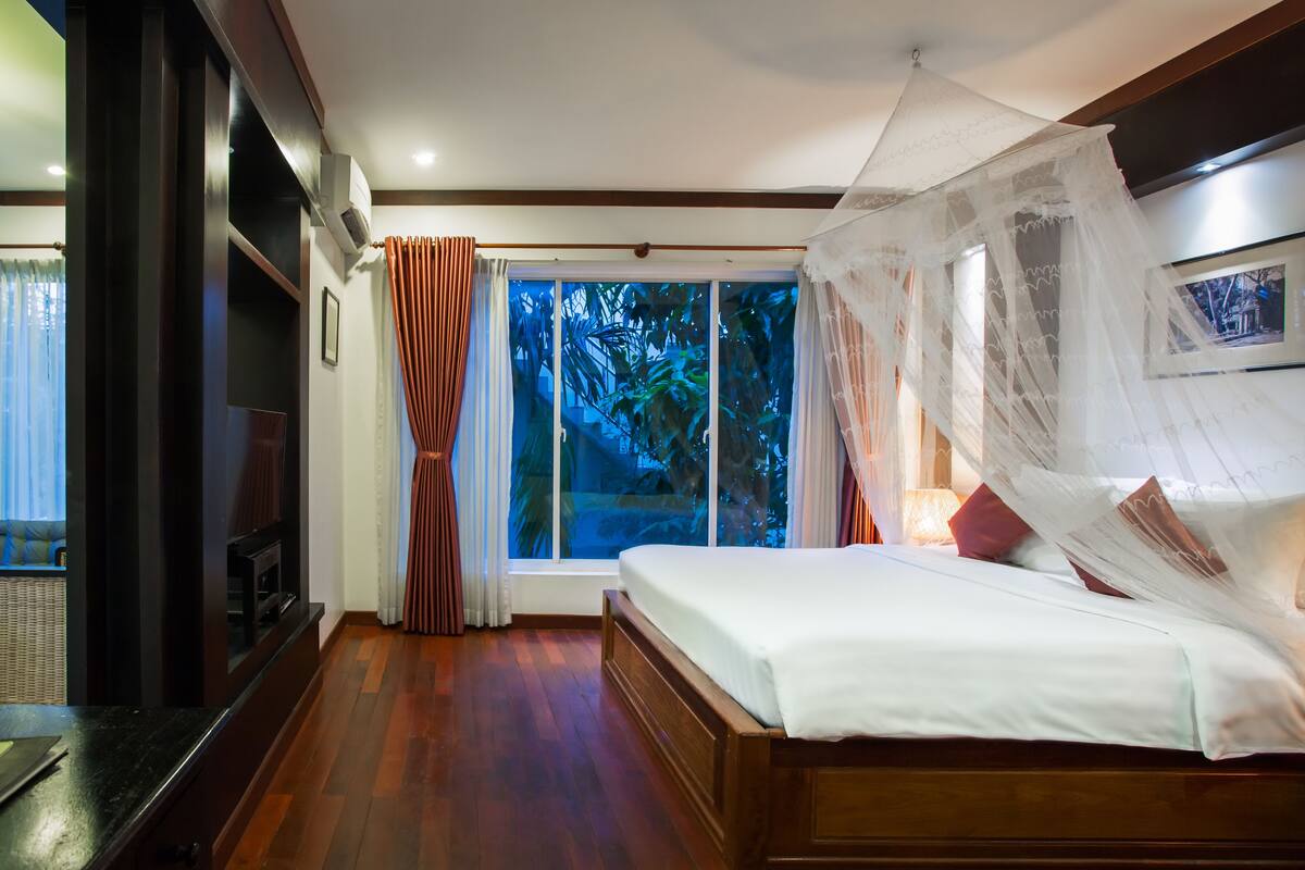 Un dormitorio con cama, televisor, ventana con cortinas y vista a árboles desde el exterior.