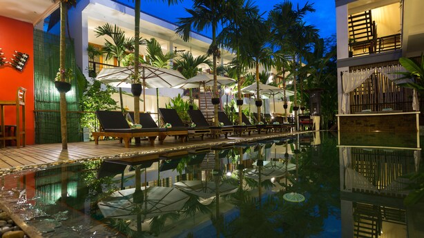 A pool area with lounge chairs and umbrellas, surrounded by palm trees and a building with multiple floors and balconies. - Diamond D'Angkor Boutique (Siem Reap)