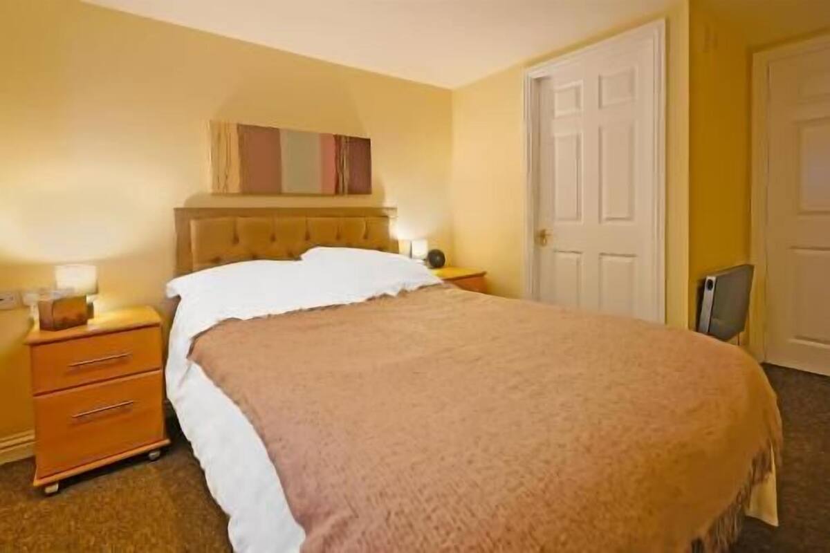 A hotel room with a bed, a nightstand, a dresser, a door, and a wall-mounted artwork.